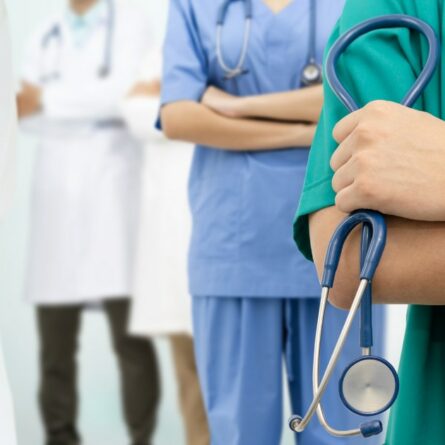 Nella foto, quattro operatori sanitari, indossando abiti da lavoro, sono in piedi con le braccia incrociate. In primo piano, una persona con uno stetoscopio appeso al collo indossa una divisa verde. Altri medici e infermieri con divise bianche e blu sfocate sono visibili sullo sfondo.